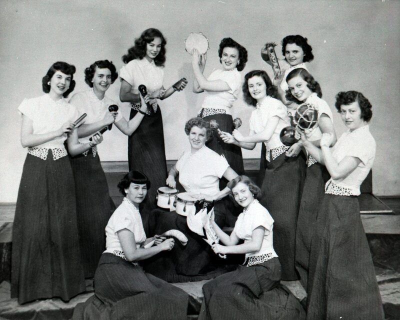 The Hormel Girls at a stage show in 1951; 11 women hold multi-cultural percussive instruments, among them bongos and a tambourine. The women wear matching ensembles of white lace trimmed blouses with long pinstriped A-line skirts. 