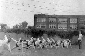 An image of Bill Zorn leading exercises on a lawn outside of Schofield Hall, 1943. Participants in the image may have been part of the 301st Army Air Forces College Training Detachment in Eau Claire.