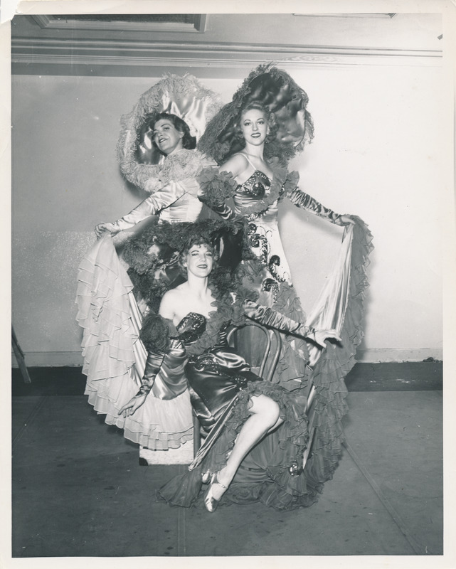 Three women posing while wearing glamourous, glittery gowns featuring ruffled edging and large cartwheel hats. The woman in the foreground is seated with legs showing. The two women in the background are standing while holding the hems of their dresses.