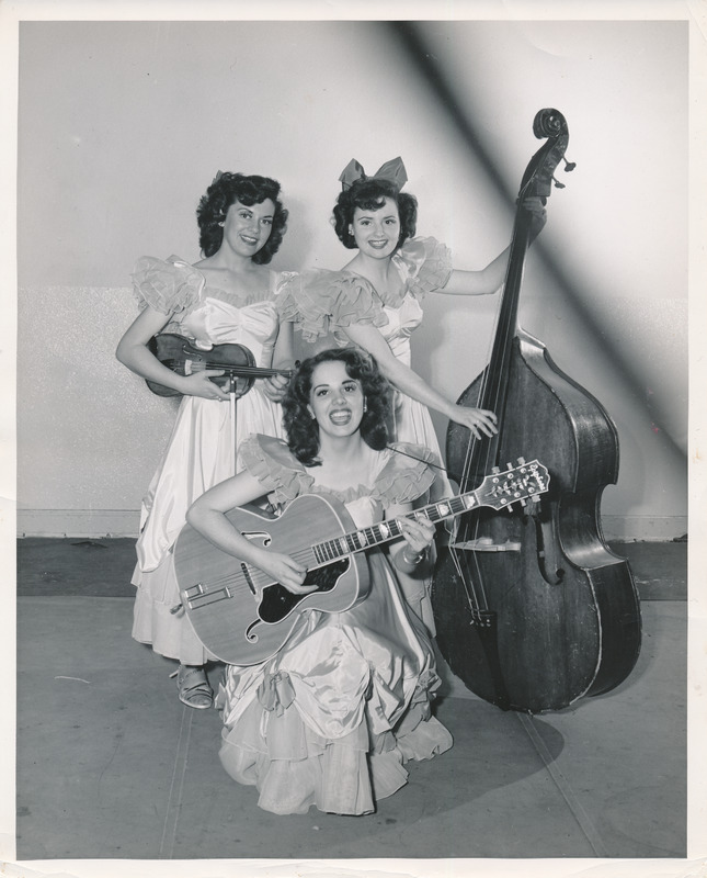 Three of the Hormel Girls pose for a photograph with their instruments; two women stand up in the back, the one on the left holds a viola, and the one on the right holds an upright bass. The woman in front is kneeling with an acoustic guitar. the women are wearing matching satin gowns with short puffy sleeves. The hems are ankle length with flower appliques and ruffles. The woman on the back right sports a large bow atop her head. 