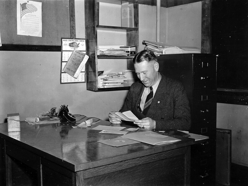 An image of Bill Zorn, wearing a suit, sitting behind a desk reviewing documents, ca. 1955.