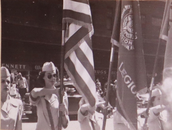 Hormel Girls stand at Attention on a city street, 3 women hold flags upright. The women wear military uniforms. 