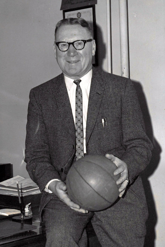 An image of coach Bill Zorn wearing a suit while sitting on a desk and holding a basketball in his hands, 1962.