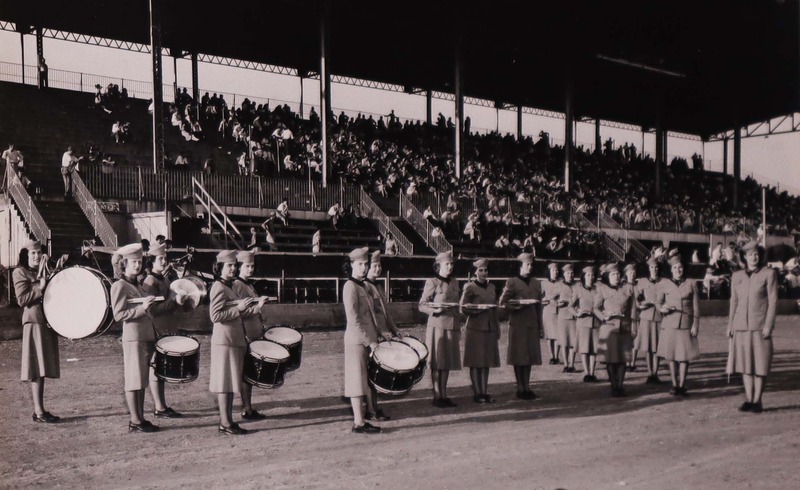 The Hormel Girls in uniform at Shreveport; 18 Hormel Girls perform for a large auditorium with a dirt floor. A sizeable audience looks on. The women wear conservative militaristic uniforms of two-piece skirt suits and felt hats. 