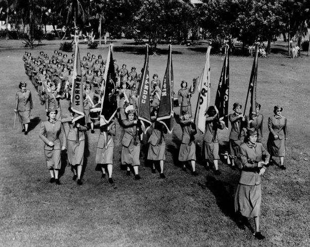 Hormel Girls march in rows in a field surrounded by trees. The women are wearing conservative military inspired skirt suits. One woman leads out front, the first row of women carries Hormel banners. Another woman observes from the side.