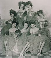 Hormel Girls Performance Group Photograph; 7 Hormel Girls pose in glamourous makeup and feather headdresses. The women wear low cut off the shoulder dresses that show off their decolletage, and full frilly skirts. The 3 women in the front row show off their bare legs and satin pumps. 